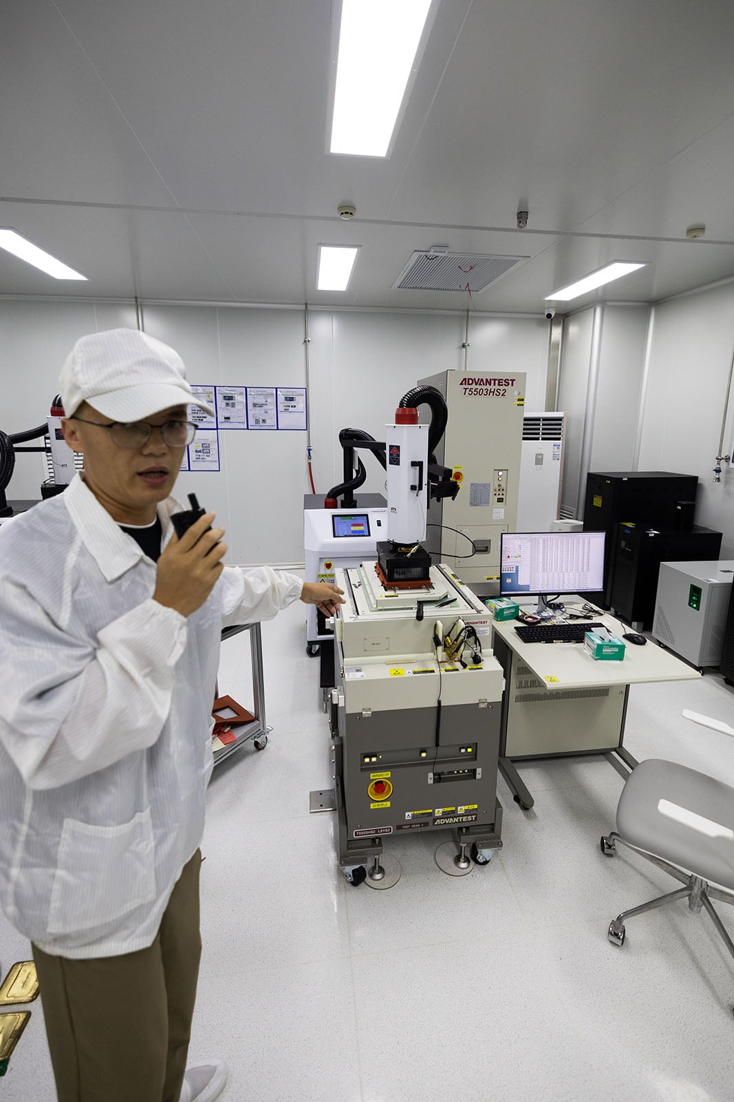 A person in a cleanroom suit and cap speaks into a handheld device, standing next to advanced laboratory equipment and computers in a well-lit, modern lab.