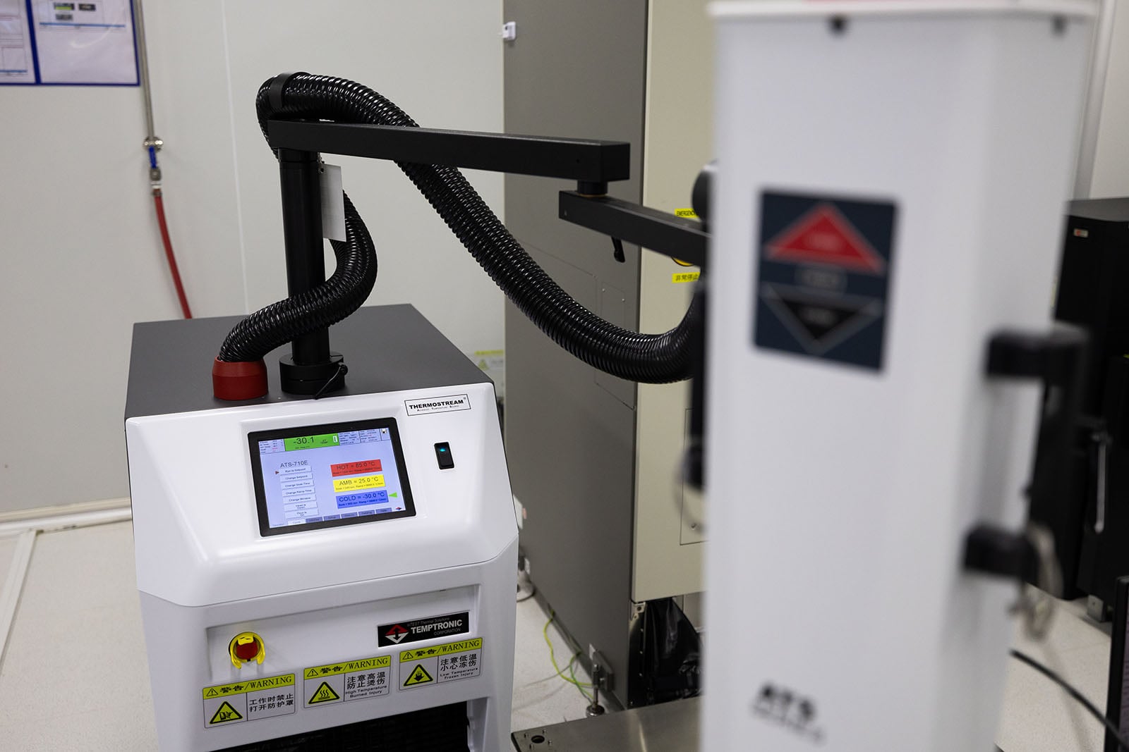 A close-up of industrial equipment in a laboratory, featuring a digital touchscreen display with warning labels, cables, and mechanical arms. The background includes a white cylindrical device with a caution sign.