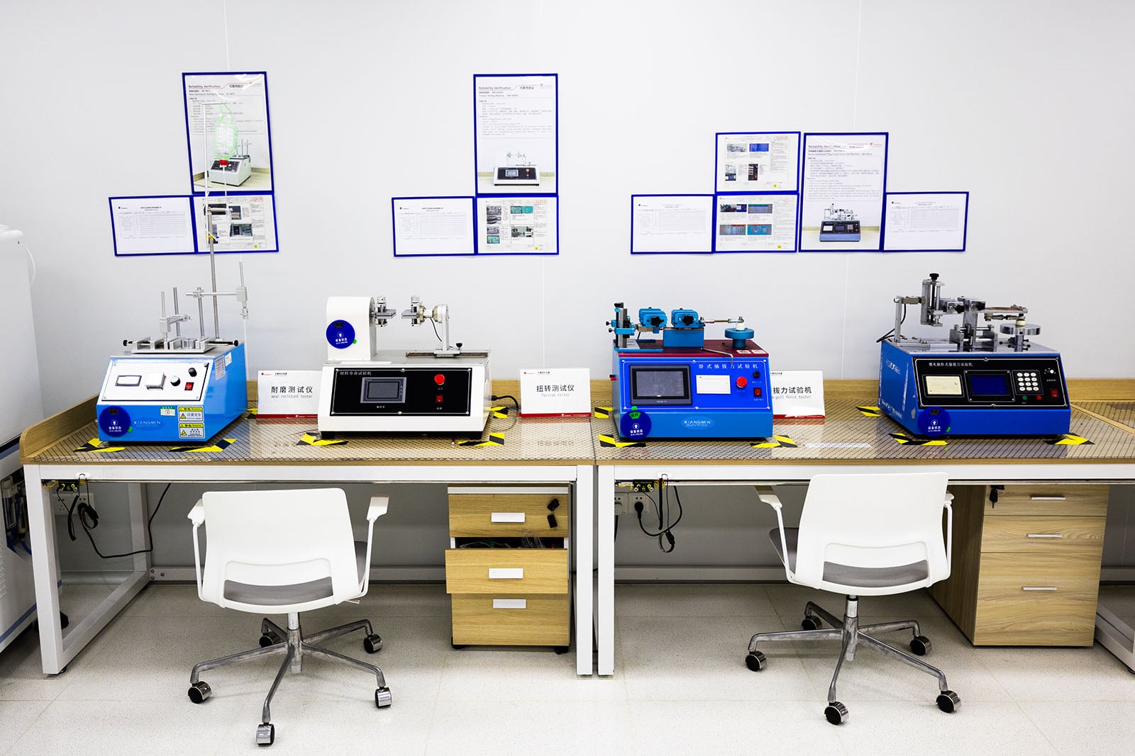 A laboratory with two desks displaying scientific equipment and machines, each with labels and digital screens. White chairs are placed at each station, and technical posters are on the wall above the desks.