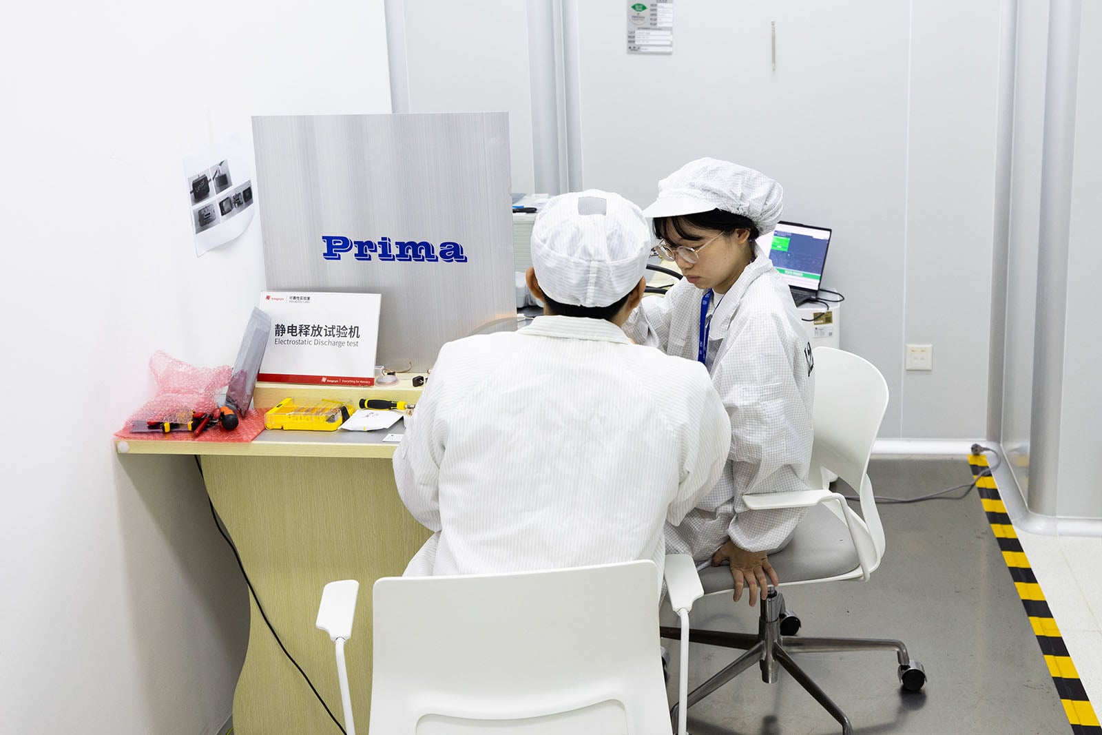 Two people in white lab coats and caps sit at a small desk in a clean, white lab room, working with computers and electronic equipment. A sign reading "Prima" is displayed on the desk.