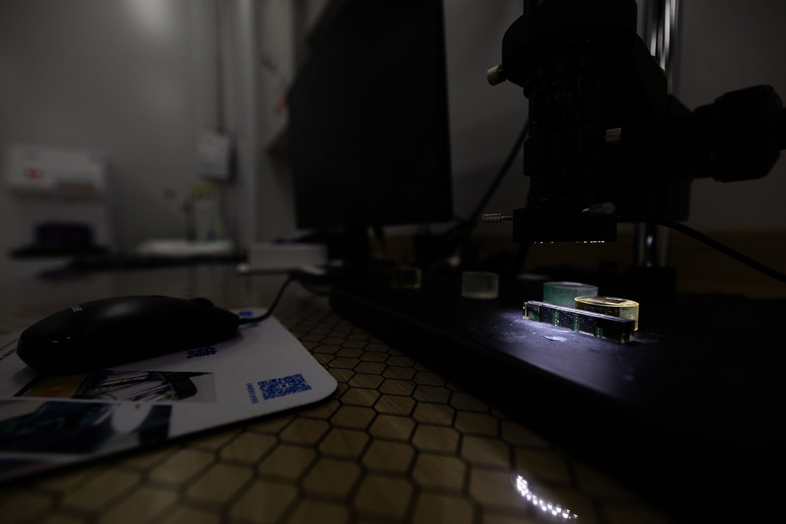 A close-up of a dark laboratory workspace featuring a microscope focused on glass slides, with a computer mouse and mouse pad nearby on a hexagonal-patterned surface.