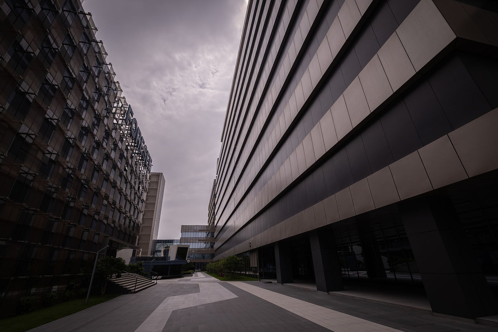 A dramatic view of modern office buildings with glass and metal facades, separated by a wide, empty walkway under a cloudy, overcast sky. The angular perspective emphasizes the height of the structures.