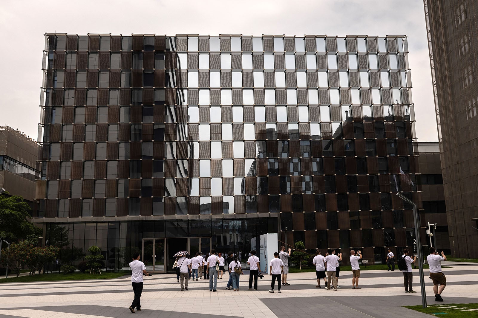 A group of people in white shirts walk toward a modern glass building with a checkered window pattern on a partly cloudy day. The courtyard is paved with geometric tiles and surrounded by greenery and other buildings.