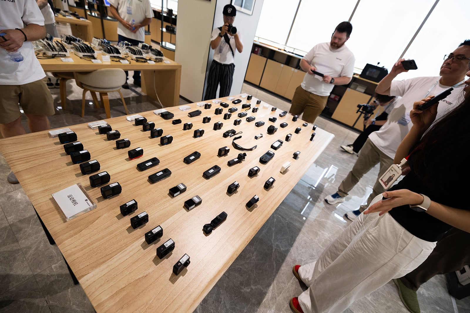 A group of people stand around a large wooden table displaying rows of small black cameras and camera lenses in a well-lit modern showroom. Some people are taking photos and observing the equipment closely.