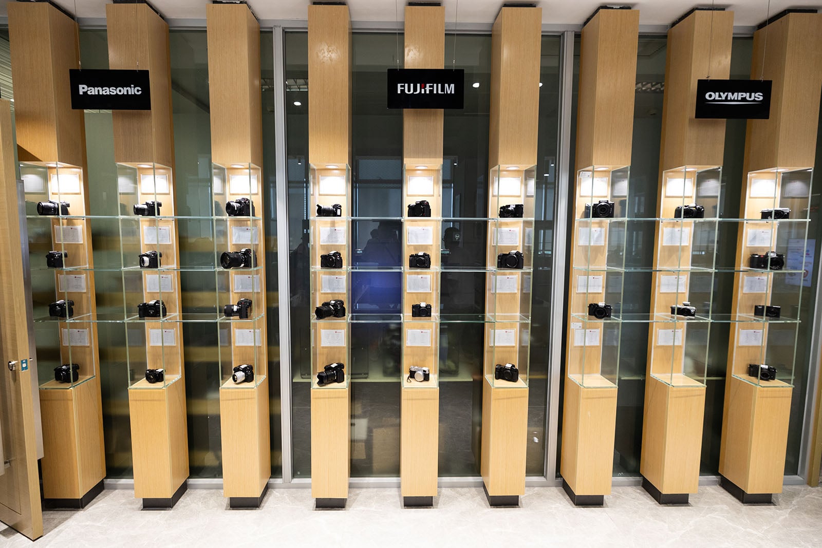  Panasonic, Fujifilm, and Olympus, in a brightly lit showroom with light wood paneling and a reflective floor.