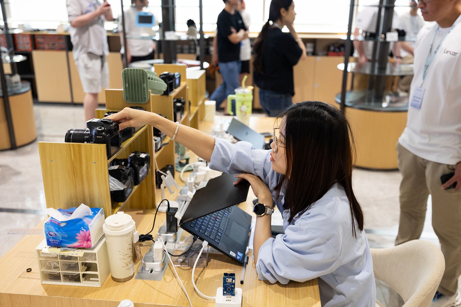 A woman sits at a desk with a laptop, reaching for a camera on a wooden shelf filled with cameras. Several people stand and talk in the background inside a modern, well-lit store or workspace.