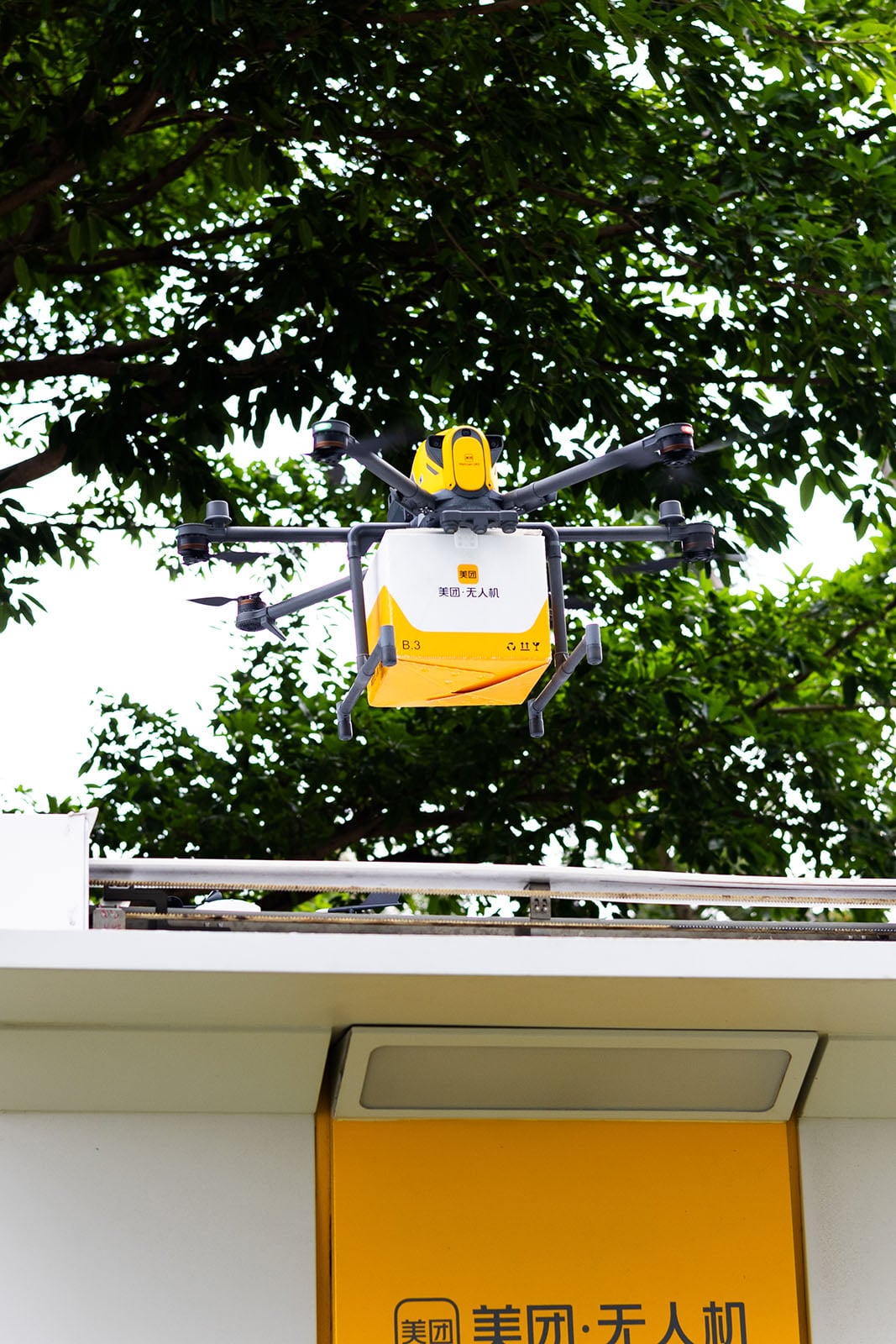 A yellow and white delivery drone hovers near a building, carrying a package, with green tree branches visible in the background.