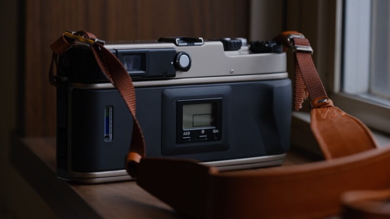 A close-up of the back of a film camera with a brown strap, resting on a wooden surface near a window, showing a small digital display and control buttons.