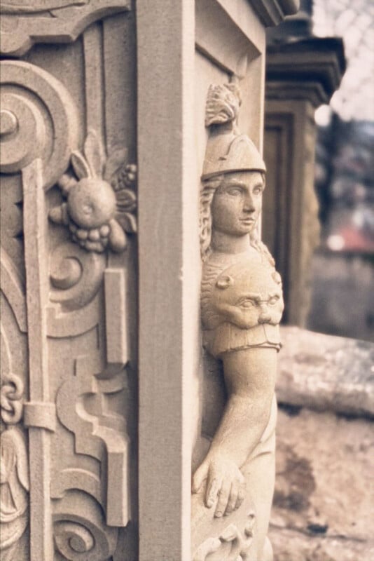 A close-up of an ornate stone sculpture shows a figure in a helmet and lion skin, carved in relief on a column with detailed floral and geometric patterns. The background is blurred.