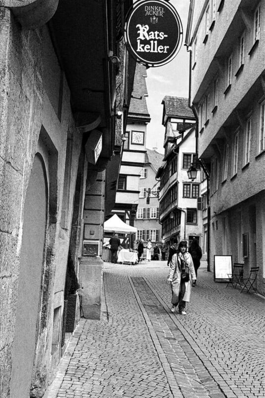 A black-and-white photo of a narrow cobblestone street lined with old buildings. Two people walk in the foreground, and a hanging sign reads "Ratskeller." More people and market stalls are visible in the distance.