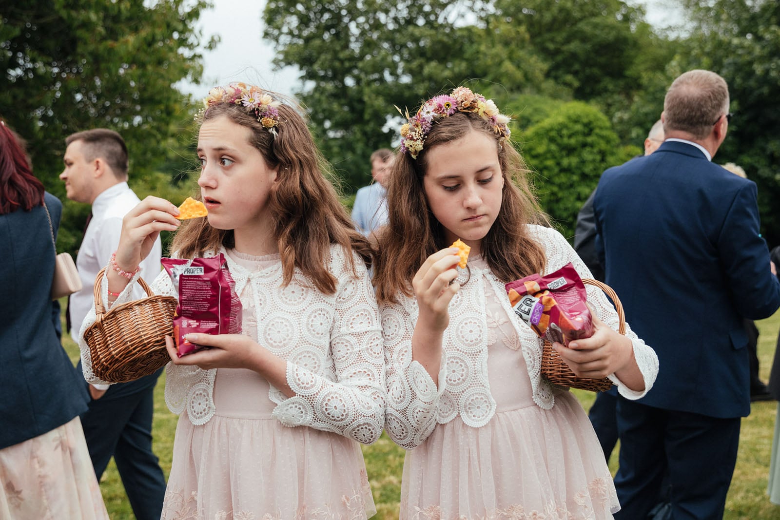 Two young girls in matching dresses and flower crowns hold baskets and snack packs while eating chips at an outdoor event, surrounded by people in formal attire.