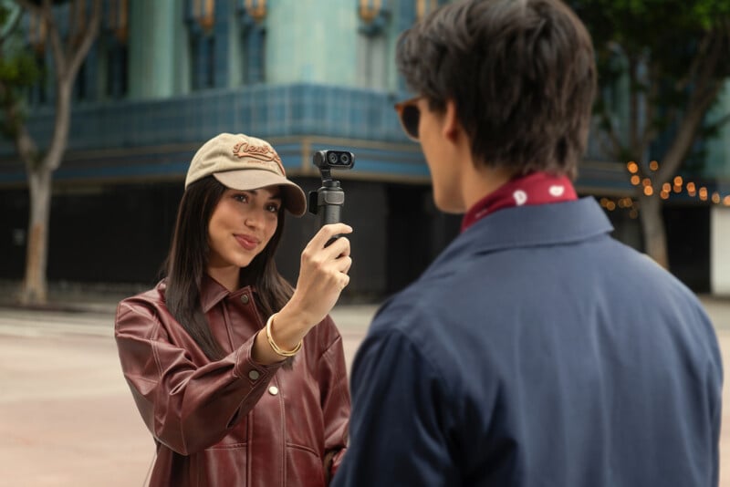 A woman in a brown jacket and beige cap is holding a small camera and filming a man in a blue jacket on a city street, with a teal building in the background.