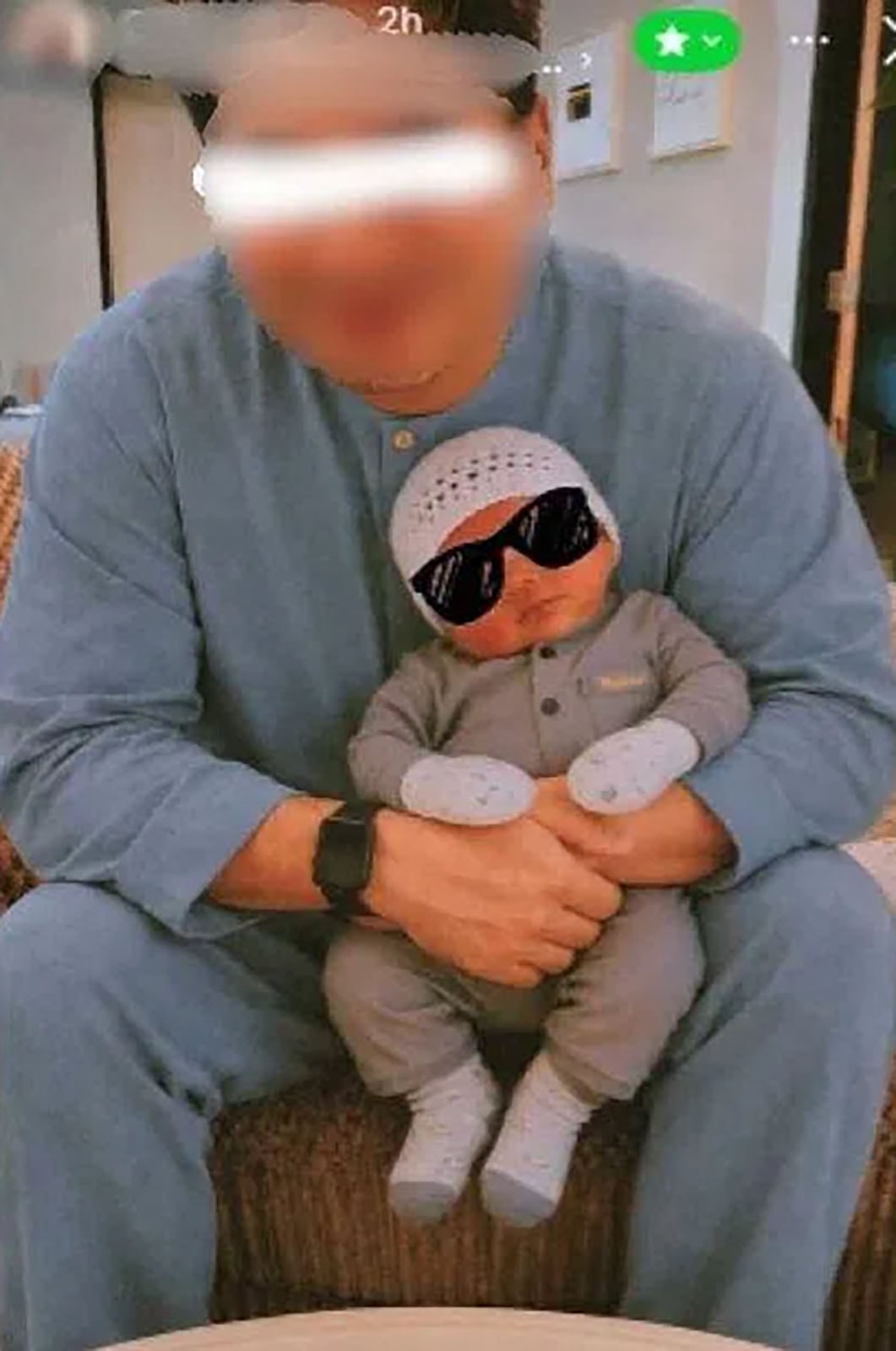 An adult wearing blue pajamas sits on a couch, holding a baby dressed in grey with a knit hat and sunglasses. Both faces are blurred. The baby’s arms are crossed and they are sitting on the adult’s lap.