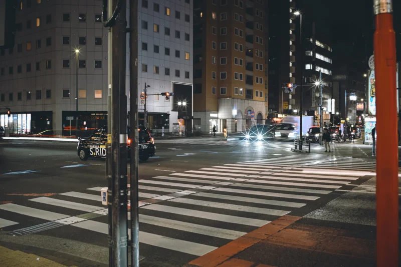 A wide, empty crosswalk at a city intersection at night, surrounded by tall buildings with lit windows. Cars with headlights on wait at the light; streetlights and traffic signals illuminate the scene.