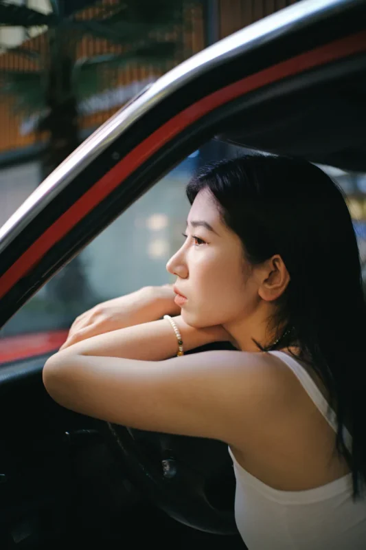 A woman in a white tank top rests her arms on the steering wheel of a car, gazing thoughtfully out of the window. Soft natural light illuminates her face.