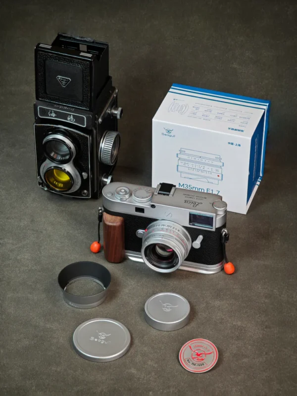 Two vintage cameras are displayed on a dark surface, one upright and one lying flat with wooden grip. Nearby are two lens caps, a lens hood, a red badge, and a blue-and-white product box labeled “M35mm F1.7.”.