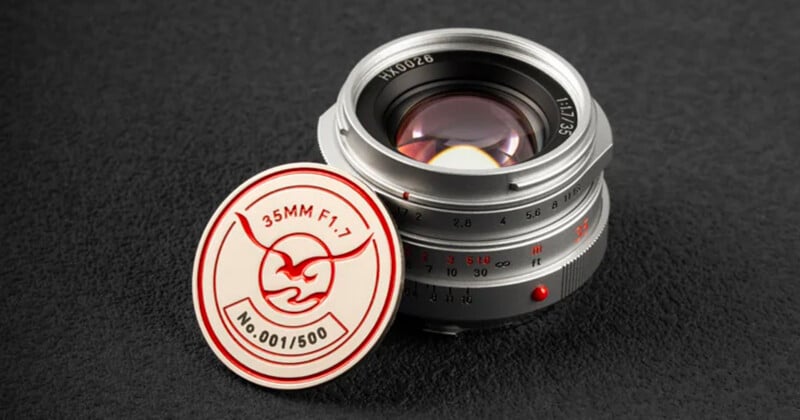 A silver camera lens labeled "35mm F1.7" sits on a textured black surface beside a round badge with a bull logo and the text "35MM F1.7 No.001/500".