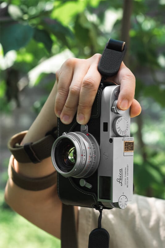 A person’s hand holding a vintage silver Leica camera outdoors, with green foliage blurred in the background. The camera is facing forward, and a black wrist strap is looped around the person's wrist.