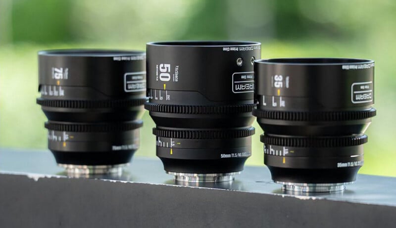 Three black camera lenses are arranged side by side on a flat surface with a blurred green background, showing their brand labels and focal lengths: 25mm, 35mm, and 50mm.