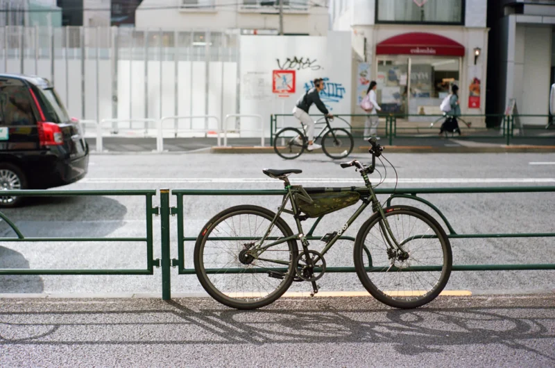 Una bicicleta está estacionada y bloqueada en una valla metálica verde junto a una calle de la ciudad. Otro hombre cruza la calle en bicicleta, con coches y peatones al fondo.