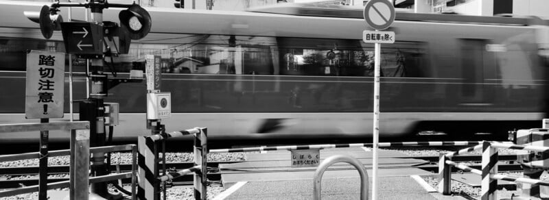 A motion-blurred train passes through a railway crossing in Japan. Warning signs, barriers, and poles are visible in the foreground. The image is in black and white.