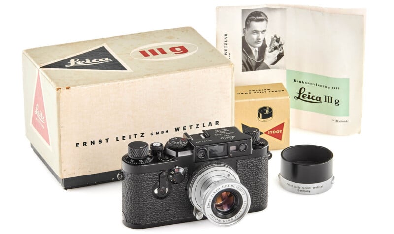 A vintage Leica IIIg camera with a lens, a lens hood, its original box, and user manuals, including one showing a man holding a camera.