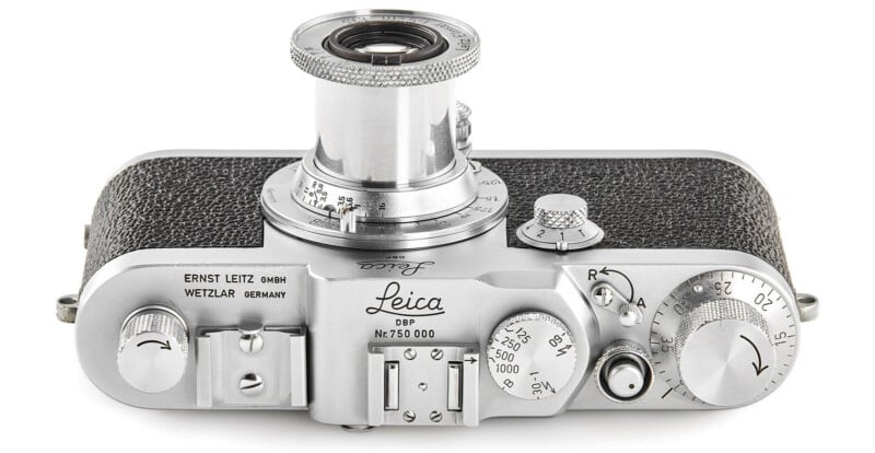 Top view of a vintage Leica film camera with a silver body, black textured grip, engraved controls, and a retractable lens. Dials, levers, and company branding are visible on the top panel.