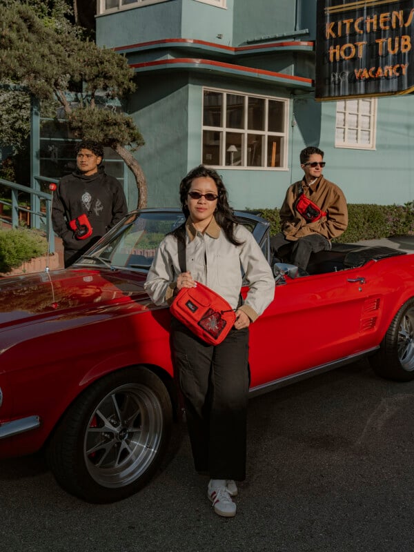 Three people pose with red bags near a red convertible parked outside a teal building. One person leans on the car while two others stand behind it. They all wear sunglasses and casual clothing.