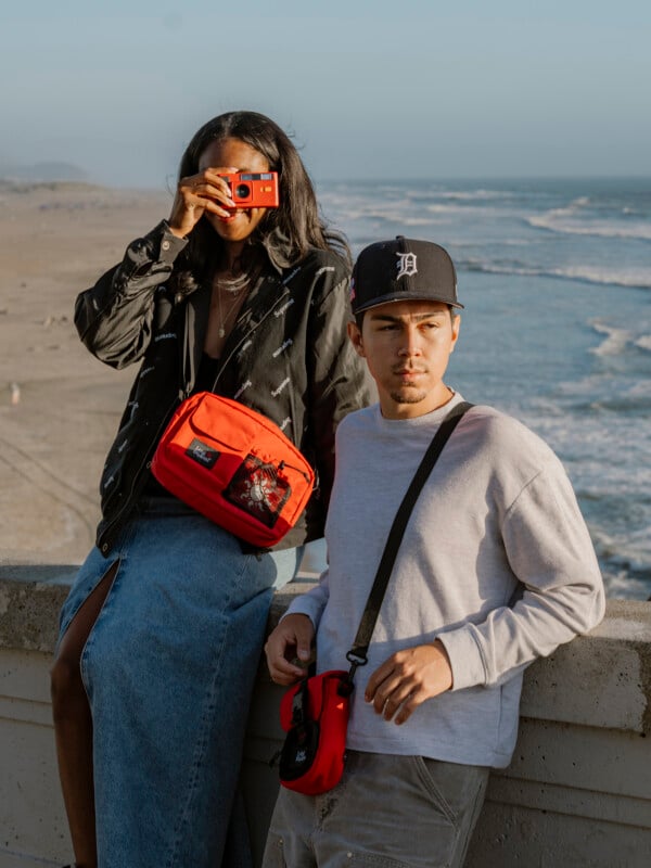 A woman holds a red camera up to her face, standing next to a man in a baseball cap, both leaning on a seaside railing with the beach and ocean in the background.