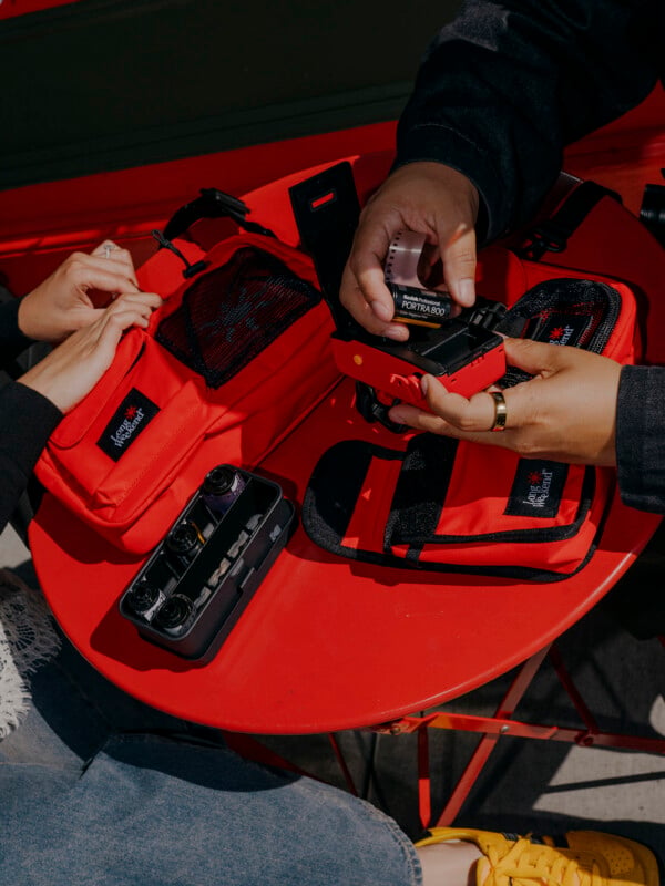 Two people sit at a red table with red camera bags, one holding a camera and a roll of film, while the other arranges items. A film camera and accessories are visible on the table.