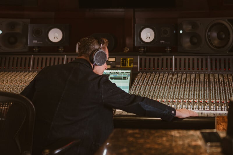 A person wearing headphones sits at a large audio mixing console in a recording studio, adjusting controls and looking at a computer screen. Multiple speakers and sound equipment are visible in the background.