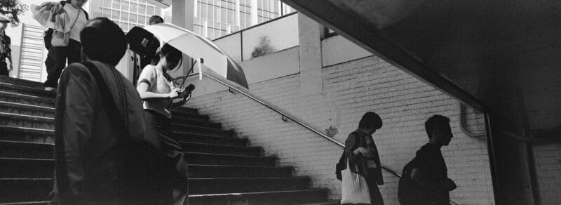 People walk up and down outdoor stairs; one person carries an umbrella and looks at a phone. The scene is in black and white, with strong light and shadow creating silhouette effects.