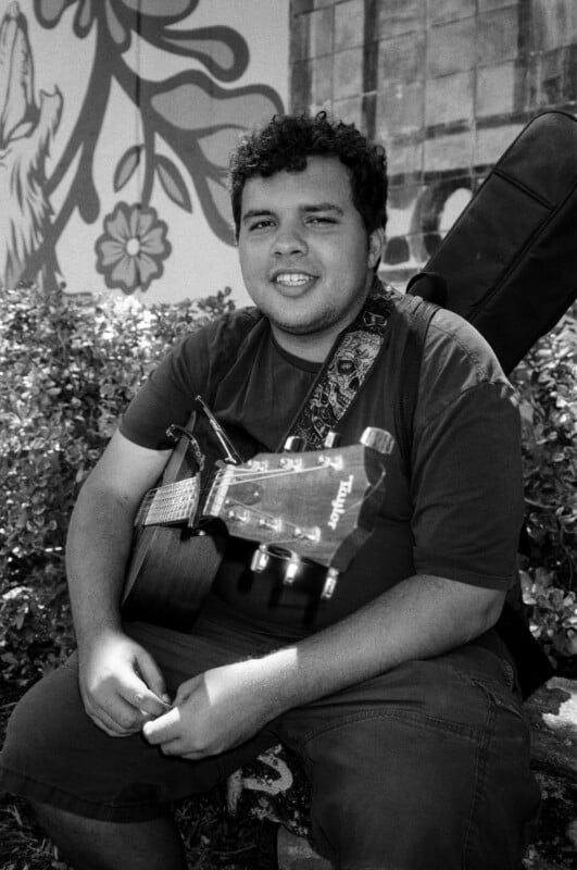 A young man sits outdoors holding an acoustic guitar, with a guitar case on his back. He is smiling at the camera. Leafy bushes and a mural with floral designs are visible in the background.