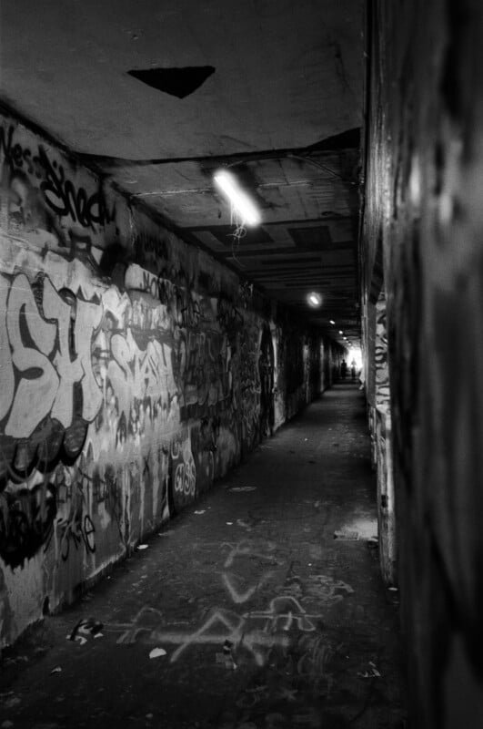A dimly lit, narrow tunnel covered in graffiti on the walls and ceiling, with a single fluorescent light overhead and a distant figure barely visible at the far end.