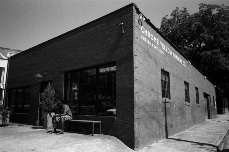 A person sits alone on a bench outside a brick building with large windows and a sign that reads "Chrome Yellow Trading Co. Coffee, Dry Goods." Trees and a street are visible around the building.