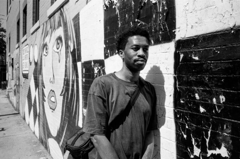 A man with a shoulder bag stands against a worn brick wall covered in graffiti art, including a large drawing of a woman's face. The photo is in black and white, creating a gritty, urban atmosphere.