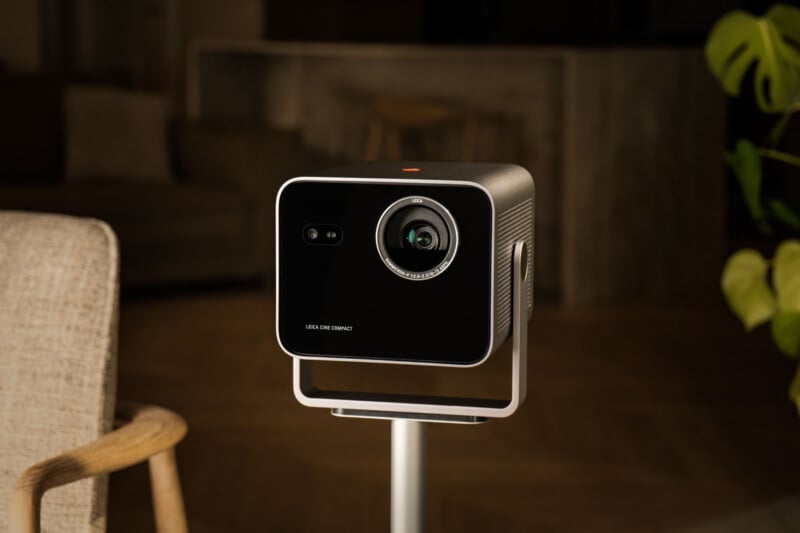 A black, cube-shaped projector with a prominent lens and a smaller sensor sits on a stand in a warmly lit room with modern furniture and plants in the background.