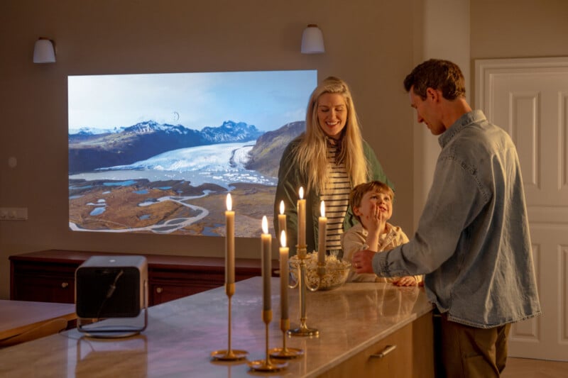 A family of three smiles in a warmly lit room, sharing popcorn at a kitchen counter with lit candles, while a scenic landscape is projected onto the wall in the background.