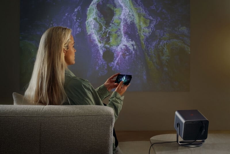 A person with long blonde hair sits on a couch, holding a smartphone while facing a projected image of colorful abstract patterns on the wall. A projector is placed on a small table beside them.