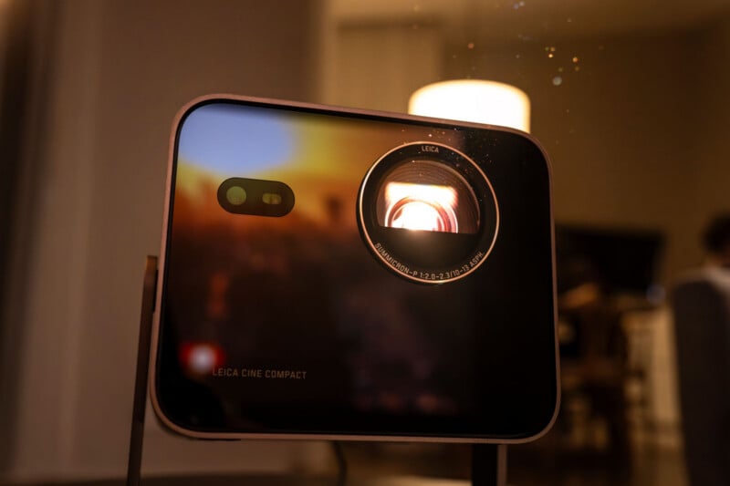 A close-up of a Leica Cine Compact projector, with its lens illuminated and projecting light. The background is softly blurred, showing a warm indoor setting with round light fixtures.