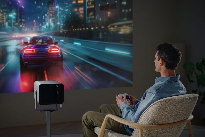 A man sits in a chair playing a racing video game projected onto a large wall screen. A projector is set up on a stand in front of him, displaying a vibrant cityscape and a red sports car on the screen.