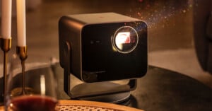 A modern black projector sits on a table, projecting colorful light beams in a dimly lit room. Candles and a woven placemat are visible nearby, creating a cozy atmosphere.