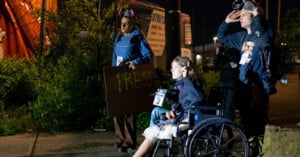 Three people are outside at night; one sits in a wheelchair holding a drink, while two others stand nearby, one holding a "PRESS" sign and the other adjusting a helmet or headgear.