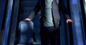A person wearing dark clothing stands on an escalator, holding a blue suitcase with one hand and resting the other hand on the escalator railing. The scene is dimly lit with blue-toned lighting.