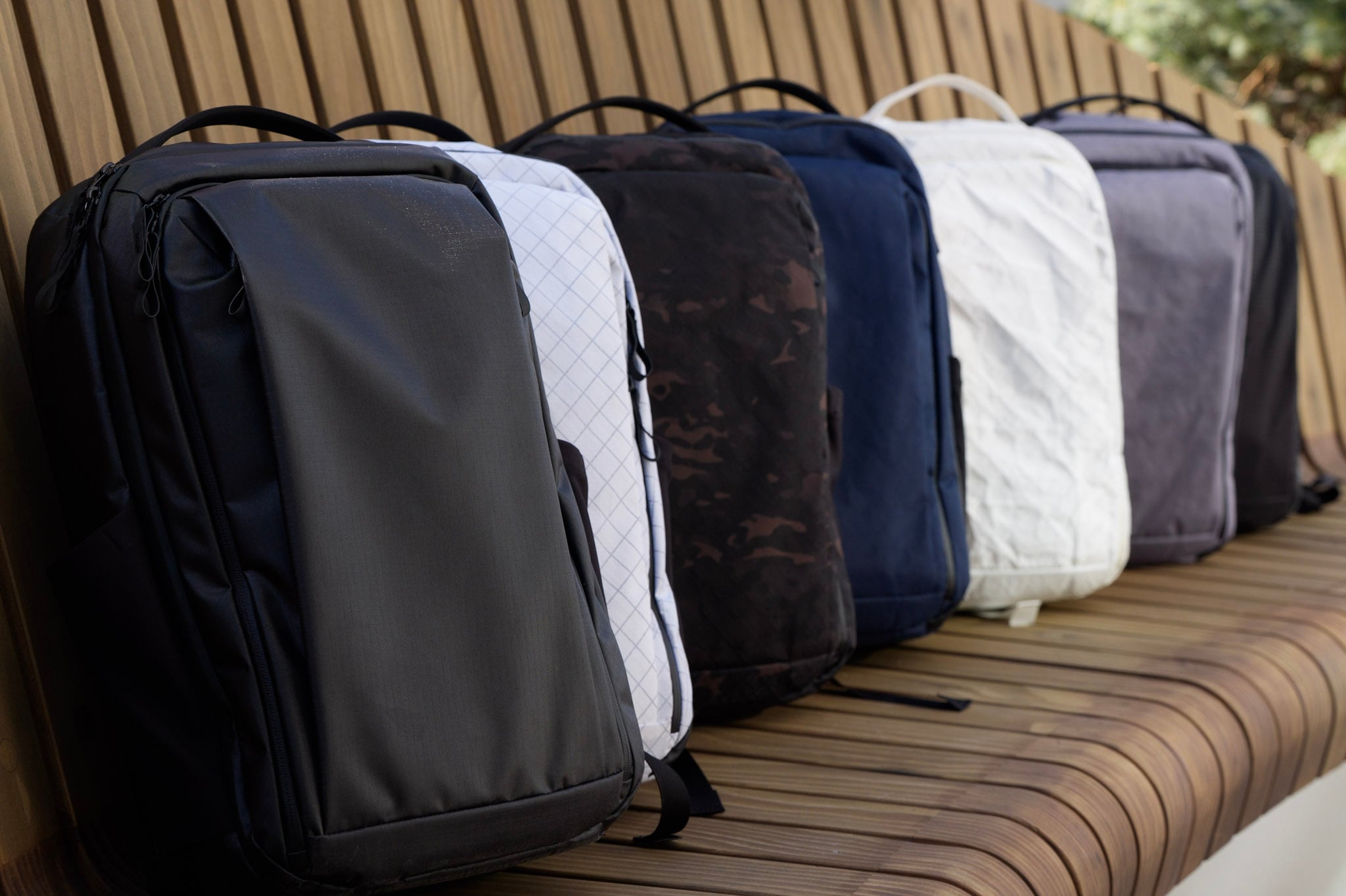 Five backpacks in different colors and patterns—black, white, camouflage, blue, and gray—are lined up on a wooden bench outdoors.
