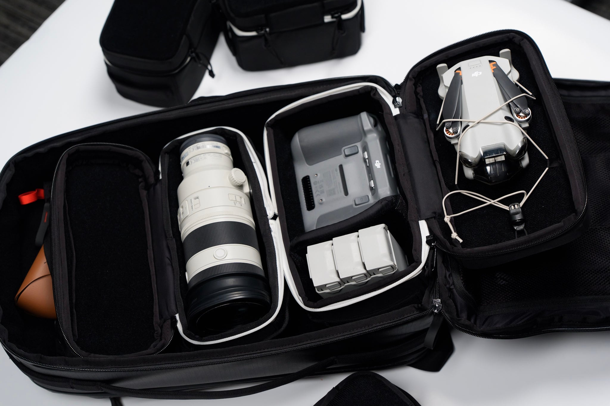 An open camera bag containing a large white camera lens, three batteries, a charging hub, and a compact drone, all neatly organized in padded compartments.