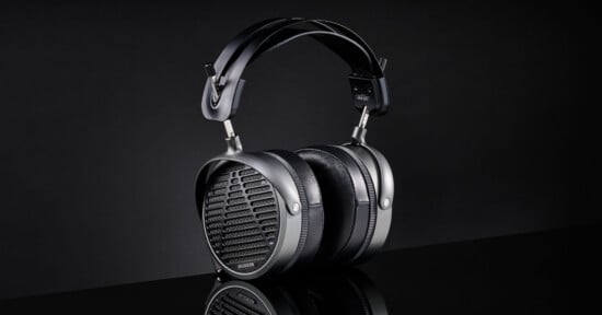 A pair of large, black over-ear headphones with a padded headband and oval-shaped earcups featuring a grille design, displayed against a dark background.