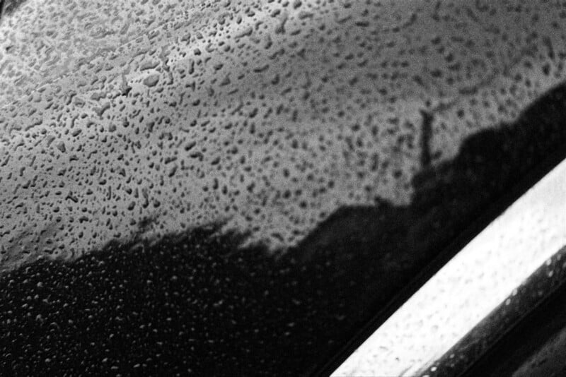 Close-up of raindrops on a reflective dark surface, possibly a car window or body, with blurred reflections of buildings and a bright diagonal metallic edge in the lower right corner. The image is in black and white.