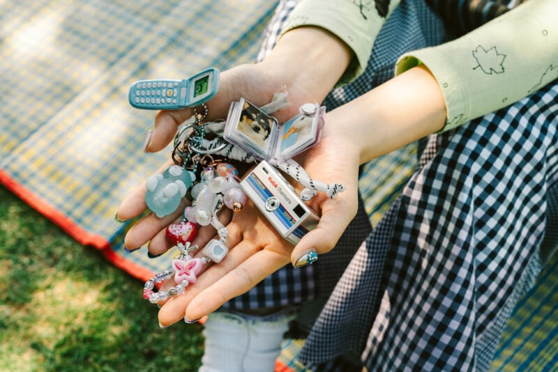 A person in checkered pants and a light green long-sleeve shirt holds several small keychain trinkets, including a toy cell phone, camera, book, beads, and charms, while sitting on a plaid blanket outdoors.