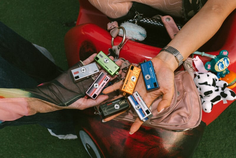 Two hands hold several colorful cassette tape keychains above a pink purse and other accessories, including a plush toy and a toy car, on a grassy surface.